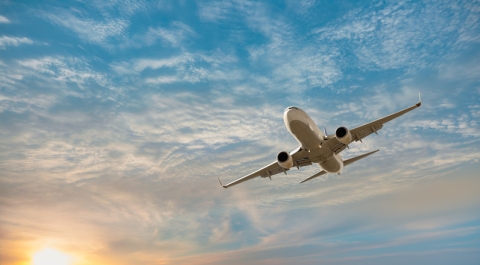 a plane glides in a shispy cloud sky with the sun dipping in the bottom left hand corner of the photo