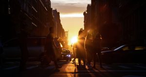 Image shows a crowded intersection with the silhouette of cars and  pedestrians crossing, while the sun sets in the background.